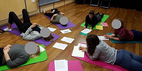 sechs Personen liegen auf dem Boden in einem Kreis auf Matten und führen so den Unterricht